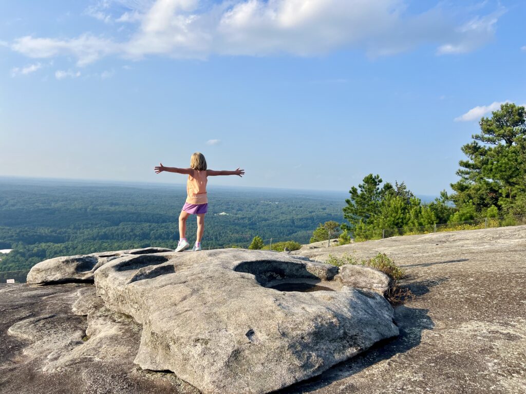 Little girl with blonde hair standing on a rock atop a mountain with her arms stretched out