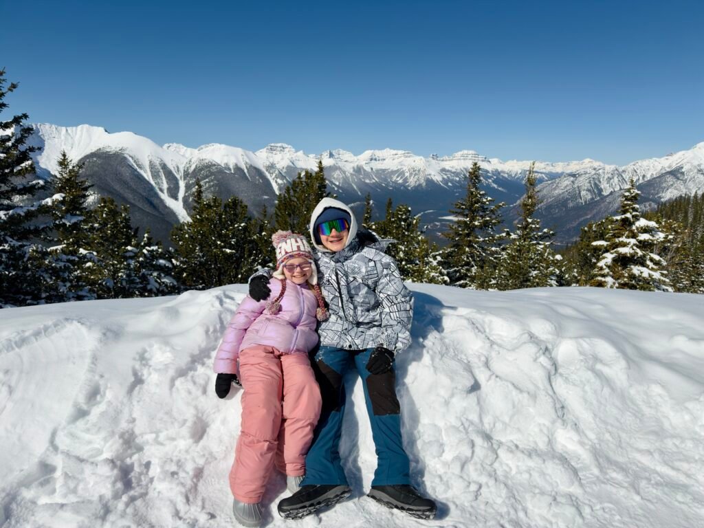 Two kids dressed in colorful winter gear sit on a snowbank at the summit of Sulphur Mountain, smiling with a panoramic view of snow-covered peaks stretching into the distance. Surrounded by frosted evergreens and bright blue skies, this scenic lookout captures a joyful moment in Banff National Park.
