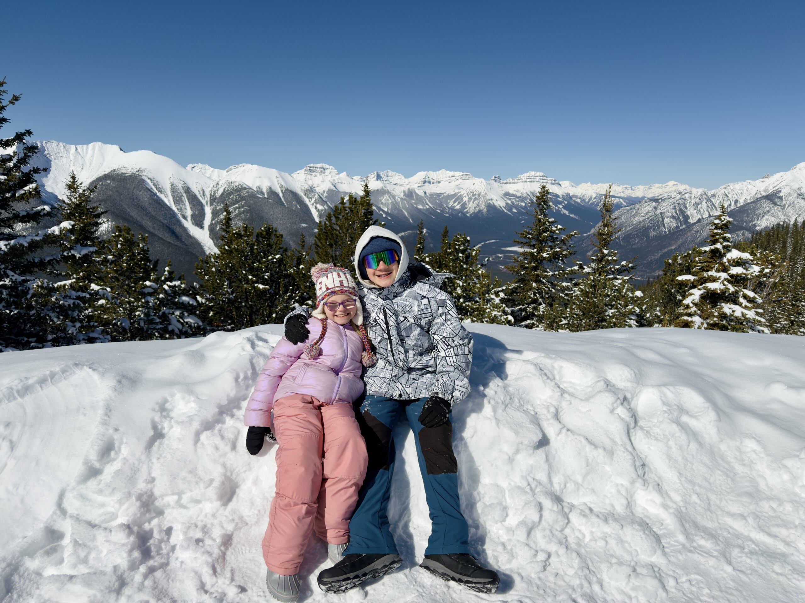 Two kids dressed in colorful winter gear sit on a snowbank at the summit of Sulphur Mountain, smiling with a panoramic view of snow-covered peaks stretching into the distance. Surrounded by frosted evergreens and bright blue skies, this scenic lookout captures a joyful moment in Banff National Park.