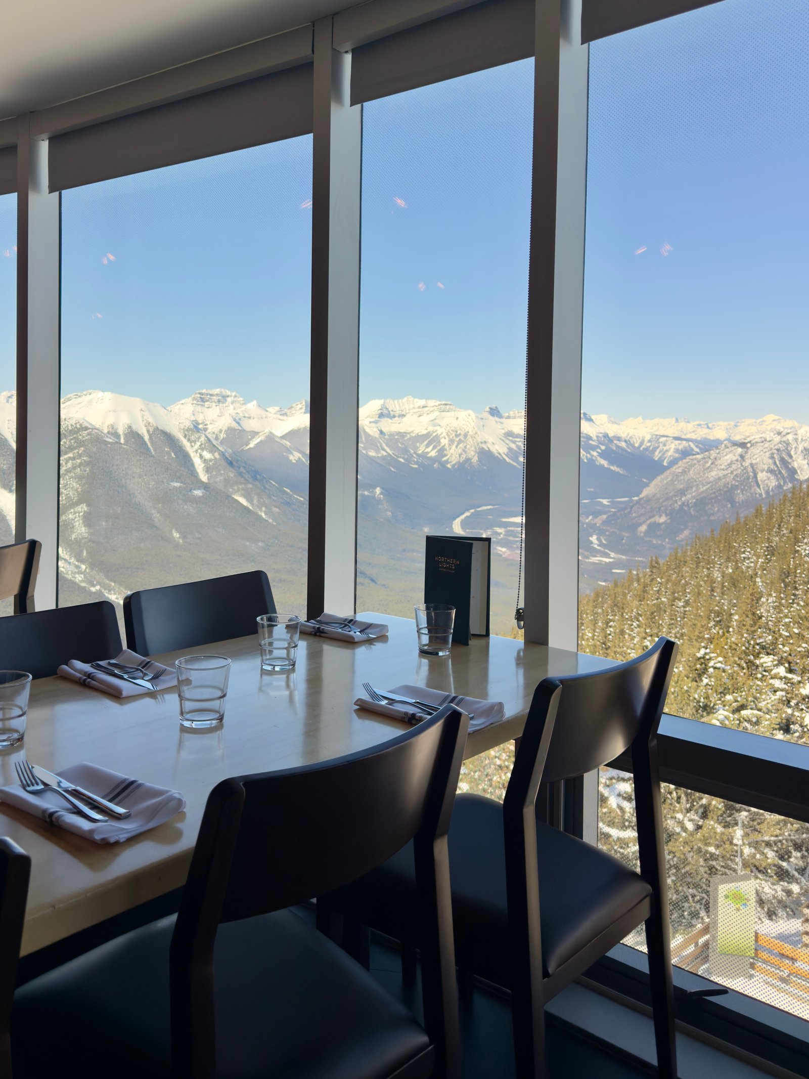 Set dining table at Northern Lights Alpine Kitchen with large windows framing a panoramic view of the snow-covered Canadian Rockies. The sunlit mountain peaks, deep valleys, and forested slopes create a stunning backdrop for this elevated Banff dining experience.