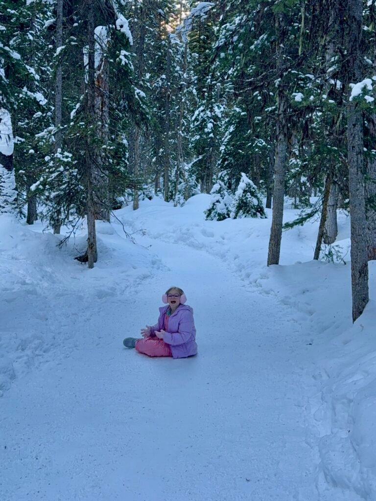 Young girl in a pink snowsuit and earmuffs sits laughing on the snow-covered Fairview Lookout Trail near Lake Louise. Surrounded by snow-dusted evergreens, the winter forest creates a magical and playful atmosphere on this family-friendly Banff hike.