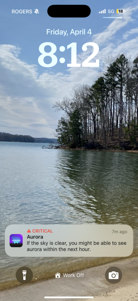 iPhone lock screen showing a lakeside view with calm water, a sandy shore, and trees under a partly cloudy sky, with a critical aurora alert notification warning that the northern lights may be visible within the hour if skies are clear.