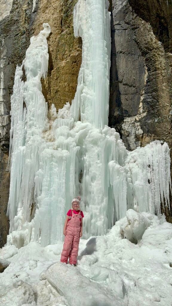 Young girl in a pink snowsuit and winter hat smiles in front of a towering frozen waterfall along the Grotto Canyon hike. The thick layers of ice cascade down a rugged rock face, creating a dramatic and icy backdrop on this winter adventure in Banff.