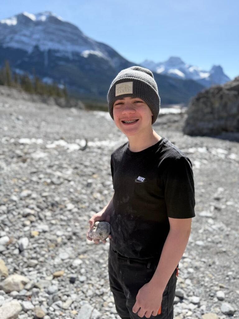 Smiling boy wearing a “Banff” beanie and black shirt holds a rock while standing on the rocky riverbed of Grotto Canyon. Towering mountains with snow-dusted peaks rise in the background under a bright blue sky, showcasing a sunny day on this scenic Banff family hike.