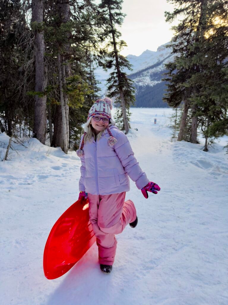 Young girl in a pink snowsuit and winter hat smiles while holding a bright red sled on a snowy path lined with pine trees. The frozen lake and distant mountain peaks glow in the soft sunset light, capturing a playful winter moment in Banff.