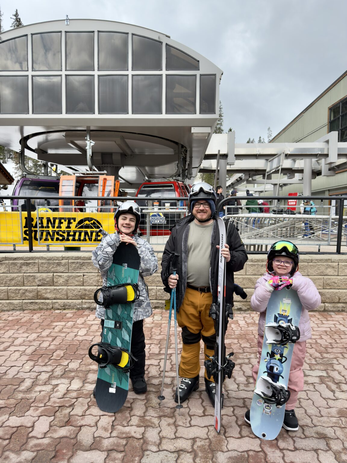 A man with skis and two kids holding snowboards smile in front of the gondola station at Sunshine Village Banff. The kids' colorful gear and excited expressions highlight a fun family day of snowboarding in Banff with kids.