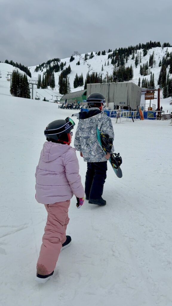 Two kids in snow gear and helmets walk toward the ski lifts at Sunshine Village Banff Ski Resort, one carrying a snowboard. Snow-covered slopes and evergreen trees frame the wintry scene, capturing a family-friendly moment of snowboarding in Banff with kids.