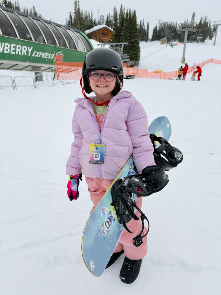 Young girl in a pink snowsuit and helmet smiles while holding a colorful snowboard at Sunshine Village ski resort. She stands in front of the Strawberry Express lift with snow-covered slopes, ski instructors, and evergreen trees in the background during a family snowboarding day in Banff.