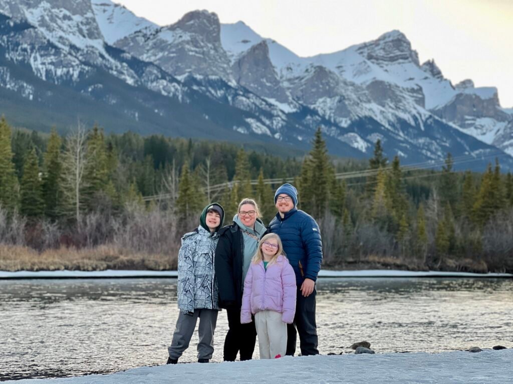 A smiling family of four poses on a snowy riverbank with the Three Sisters mountain peaks towering in the background near Canmore. Evergreen trees and a calm, partially frozen river reflect the crisp winter setting, making this a scenic stop on a Banff family itinerary.