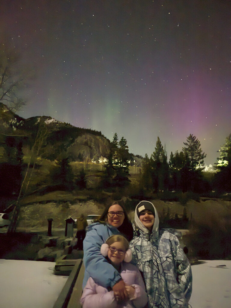 A smiling family bundled in winter coats and earmuffs poses under a starry sky with faint green and purple northern lights above snow-dusted hills and pine trees in Banff.