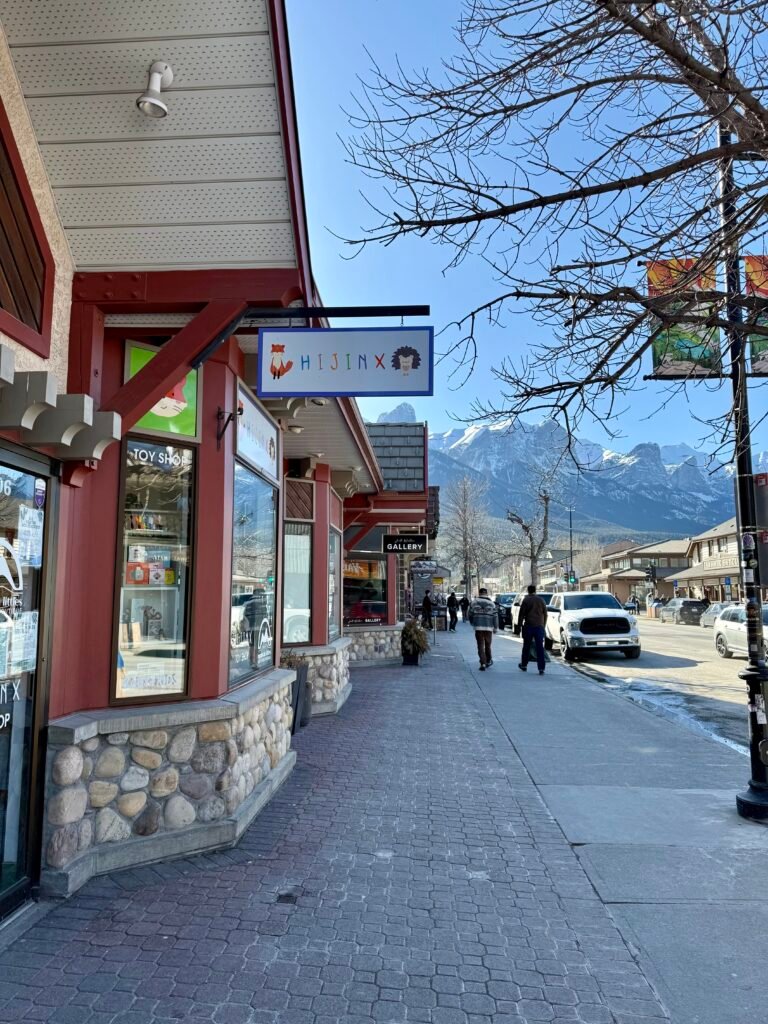 Sidewalk view of downtown Canmore featuring Hijinx Toy Shop, with its playful fox and hedgehog sign hanging above the entrance. The shop’s large windows display colorful toys and souvenirs, set against a backdrop of snowy mountain peaks under a clear blue sky.