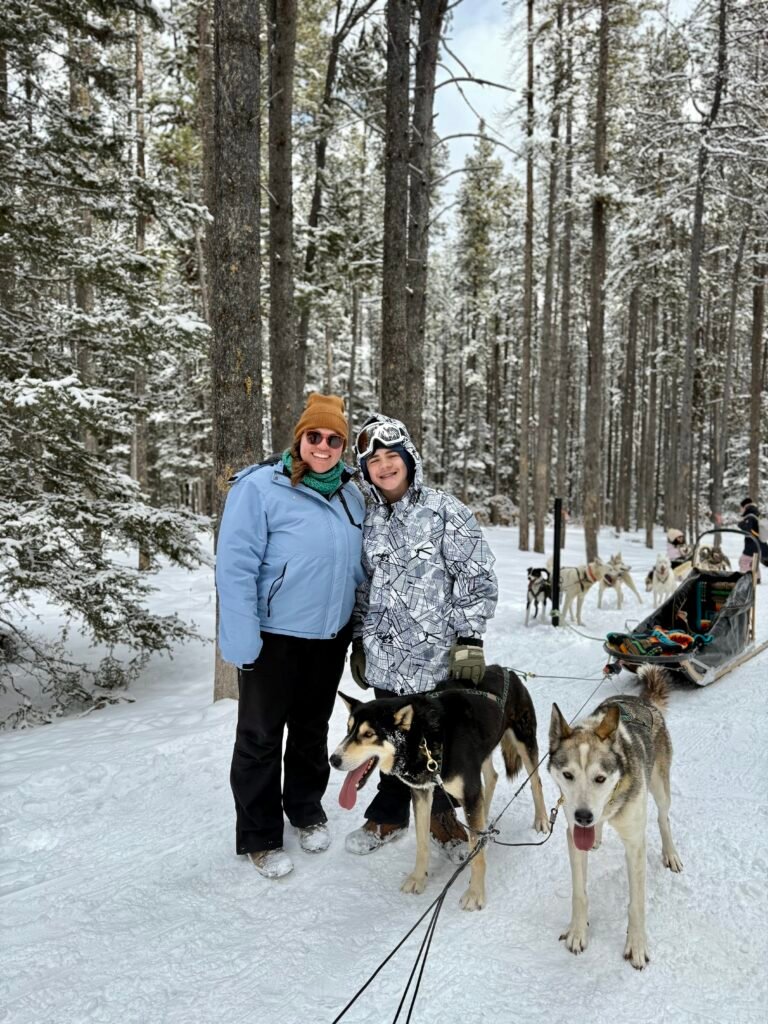 Two people bundled in winter gear stand smiling beside two sled dogs on a snowy forest trail in Banff. A traditional dog sled with a team of huskies is visible in the background, set against tall pine trees dusted with snow. This image captures a moment from a family-friendly sled dog tour, part of a Banff family itinerary.