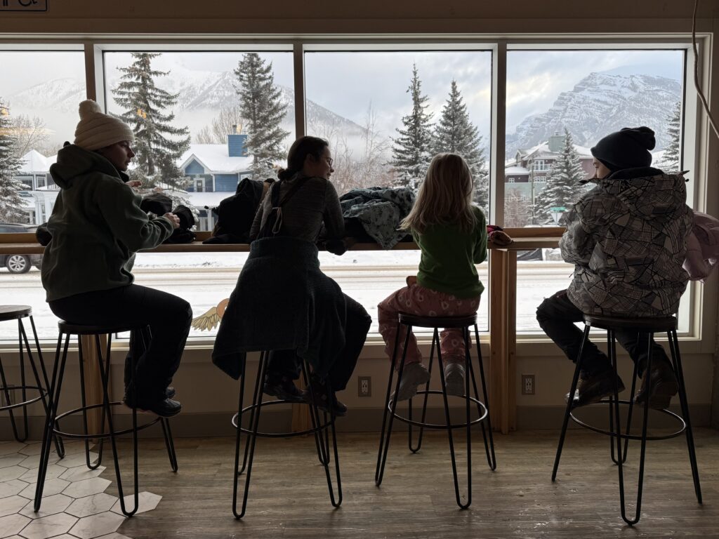 Four people, including two children, sit on stools by a large window inside Rocky Mountain Bagel Co. in Canmore. Snow-covered trees and mountains are visible outside, creating a cozy and family-friendly atmosphere typical of kid-friendly restaurants in Canmore.