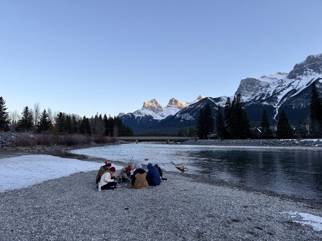 A group of families enjoys a winter picnic on the snowy banks of the Bow River in Canmore, sharing takeout from Marra’s Way Sushi. The Three Sisters peaks rise in the background, capturing the blend of scenic views and local eats that make kid friendly restaurants in Canmore memorable.