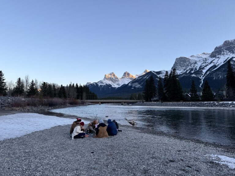 A group of families enjoys a winter picnic on the snowy banks of the Bow River in Canmore, sharing takeout from Marra’s Way Sushi. The Three Sisters peaks rise in the background, capturing the blend of scenic views and local eats that make kid friendly restaurants in Canmore memorable.