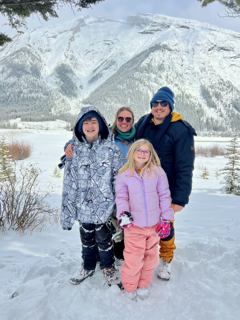 Family bundled in colorful winter gear smiles in front of a snow-covered mountain range in Kananaskis, Alberta. The scene captures a cheerful moment during a snowy day trip, ideal for showcasing a winter adventure in Banff on a budget.