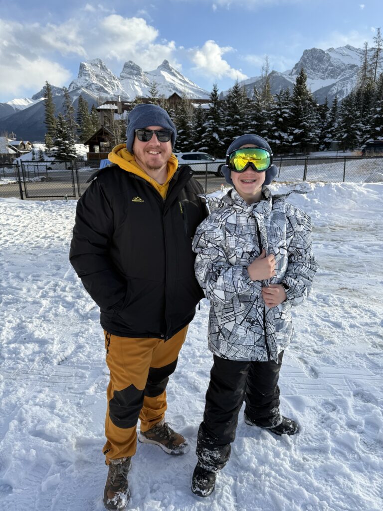 Two people dressed in insulated winter gear stand on a snowy path with the iconic Three Sisters mountain peaks behind them in Banff. They're wearing snow boots, snow pants, ski jackets, gloves, and hats — key items to pack for Banff to stay warm and comfortable during outdoor adventures.