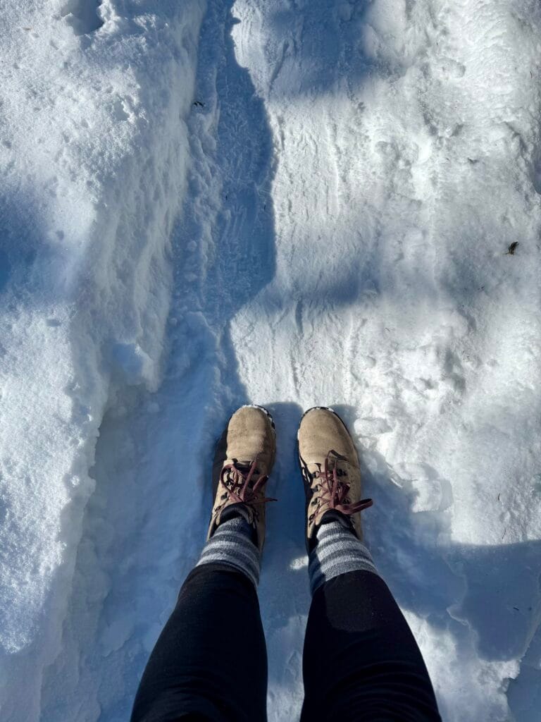 A person wearing Danner Women's Inquire Chukka hiking boots and striped Pacas crew socks stands on a packed snowy trail surrounded by deep snowbanks. The boots are tan with burgundy laces and show slight snow wear, suggesting active use in winter conditions.
