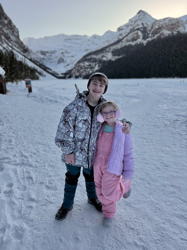 Two kids dressed in winter gear smile and pose on a snowy path surrounded by towering, snow-covered mountains in Banff. The older child wears a patterned ski jacket, snow pants, and boots, while the younger child is bundled in pink snow overalls, a puffy jacket, and earmuffs — a cozy example of what to pack for Banff in winter.