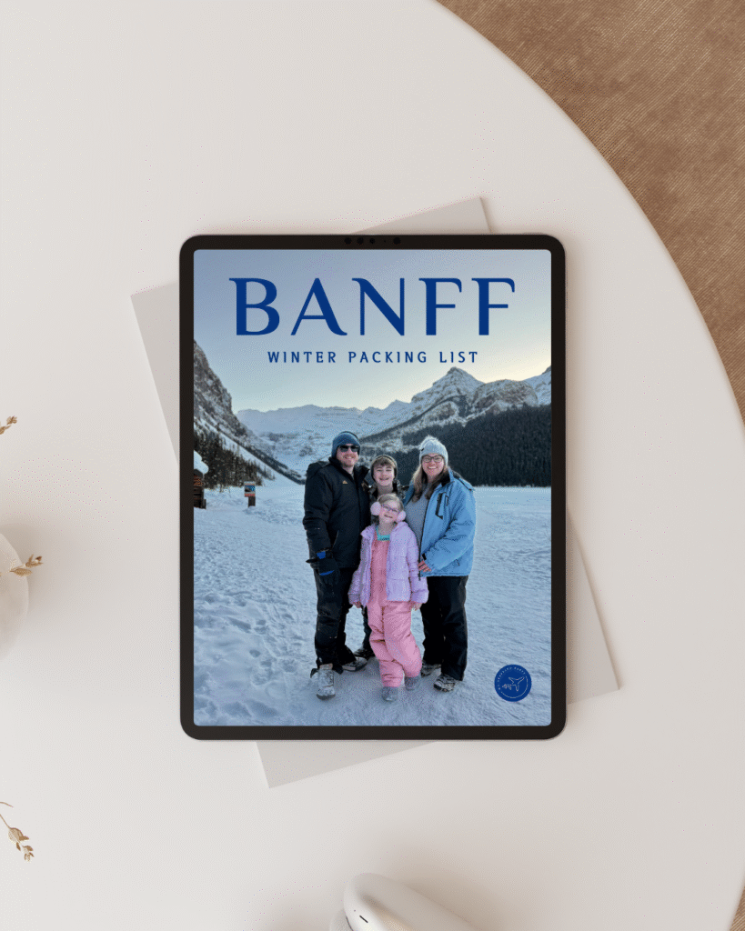 A tablet on a white table displays a digital cover titled “Banff Winter Packing List” with a family of four dressed in winter gear standing on a snowy path with snow-covered mountains and pine trees in the background. The design suggests a free downloadable guide for planning a winter trip to Banff.