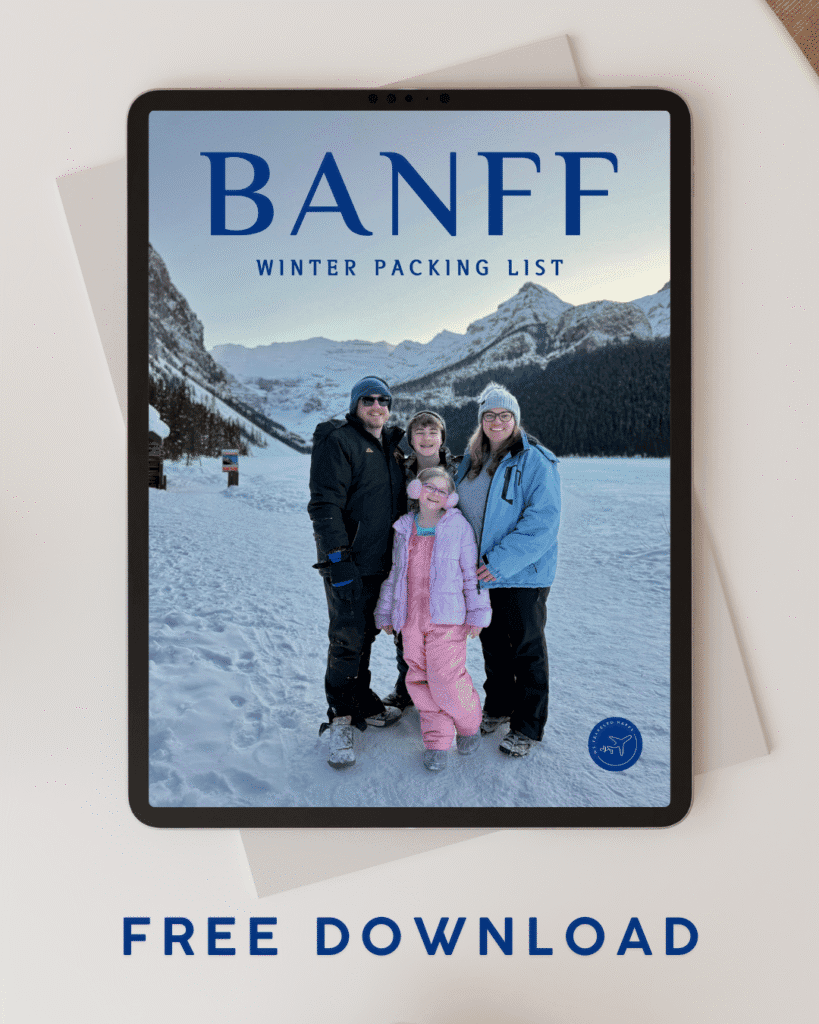 A tablet on a white table displays a digital cover titled “Banff Winter Packing List” with a family of four dressed in winter gear standing on a snowy path with snow-covered mountains and pine trees in the background. The design suggests a free downloadable guide for planning a winter trip to Banff.