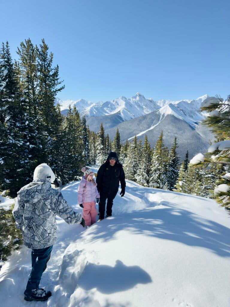 A family walks through deep snow along a scenic trail at the Sulphur Mountain summit in Banff, surrounded by snow-covered pine trees and towering Rocky Mountain peaks under a clear blue sky. A child in a pink snowsuit and winter hat smiles while walking with two adults, enjoying a sunny winter day.