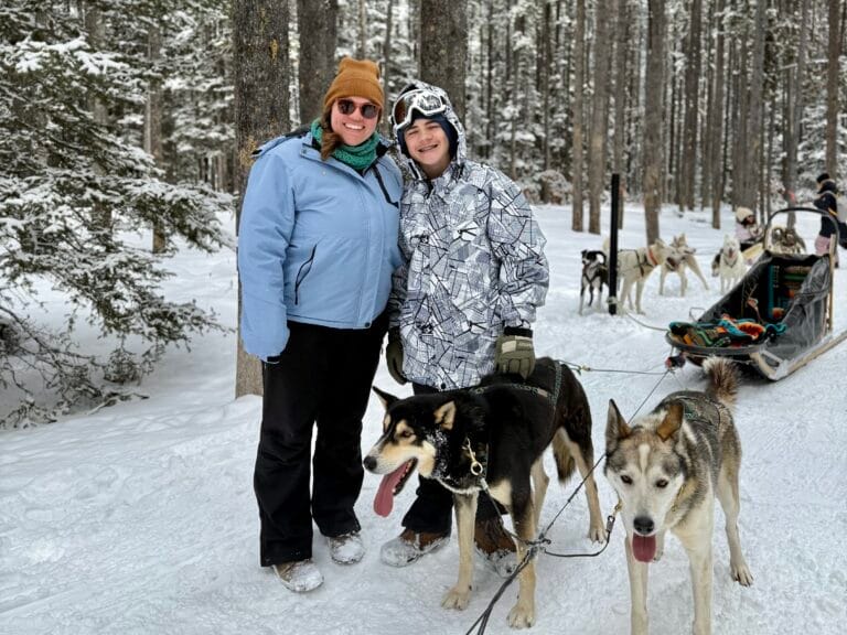 Two smiling people in winter gear pose in a snowy forest with sled dogs harnessed and ready for a tour. A traditional dog sled with colorful blankets is visible behind them, surrounded by snow-covered trees. The scene captures the atmosphere of a Snowy Owl Sled Dog Tour adventure.