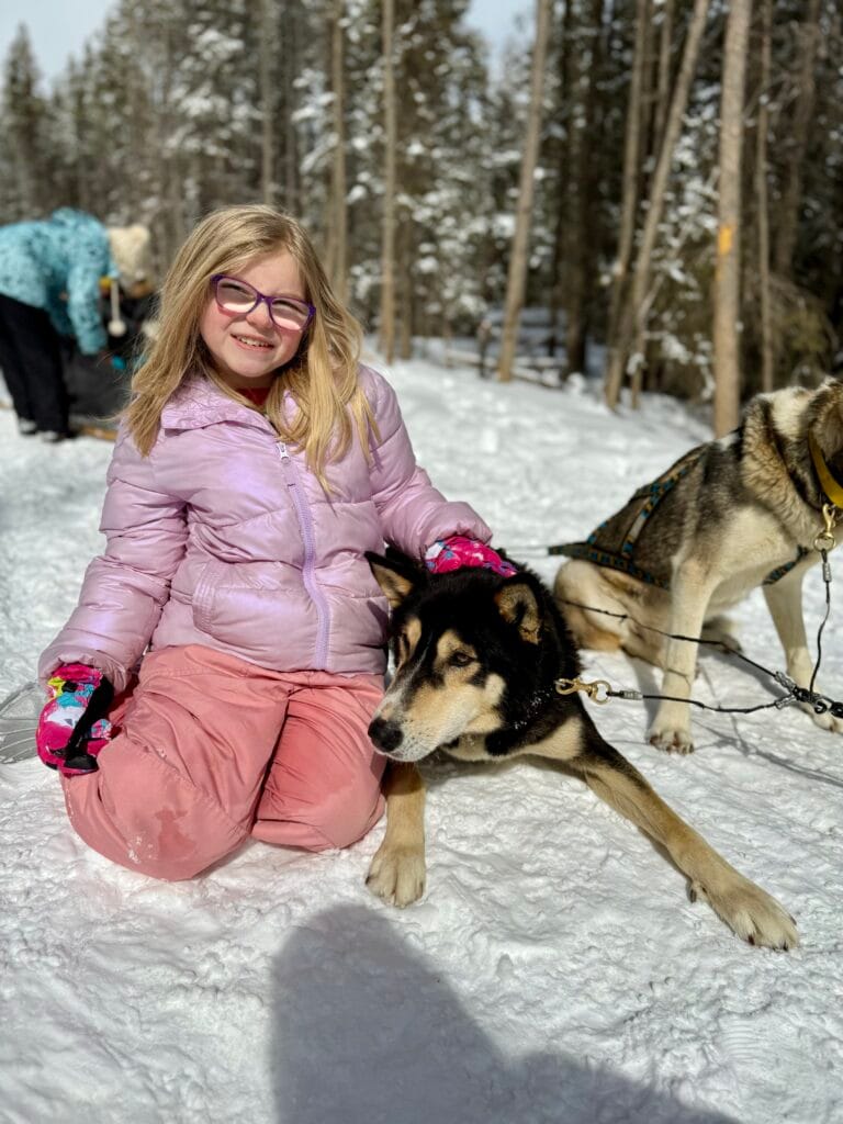 A smiling young girl in a pink jacket and snow pants kneels in the snow beside a resting sled dog during a snowy owl sled dog tour. Tall pines and another guest near a sled are visible in the wintry forest background, capturing a joyful moment that reflects positive snowy owl sled dog tours reviews.
