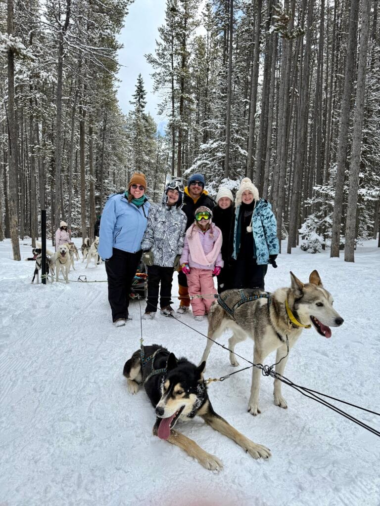 A smiling group of people poses in a snowy forest behind two huskies harnessed to a sled during a snowy owl sled dog tour. With snow-covered trees all around and happy faces, the scene reflects the family-friendly adventure often praised in snowy owl sled dog tours reviews.