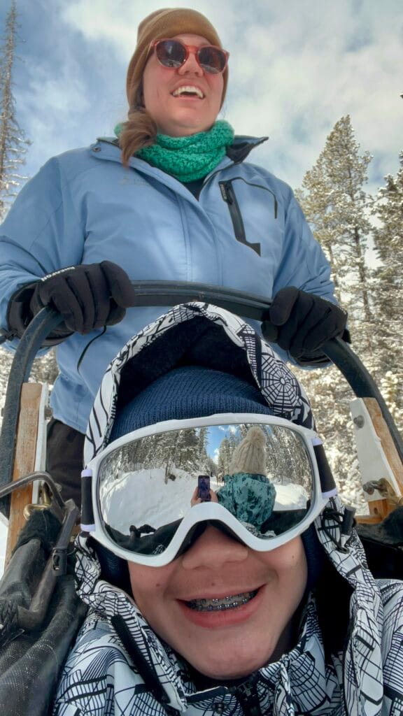 A close-up of a smiling child wearing snow goggles and braces, with the snowy forest trail reflected in the lenses during a snowy owl sled dog tour. An adult guide stands behind them on the sled, both bundled in winter gear, capturing the fun and scenic experience highlighted in snowy owl sled dog tours reviews.